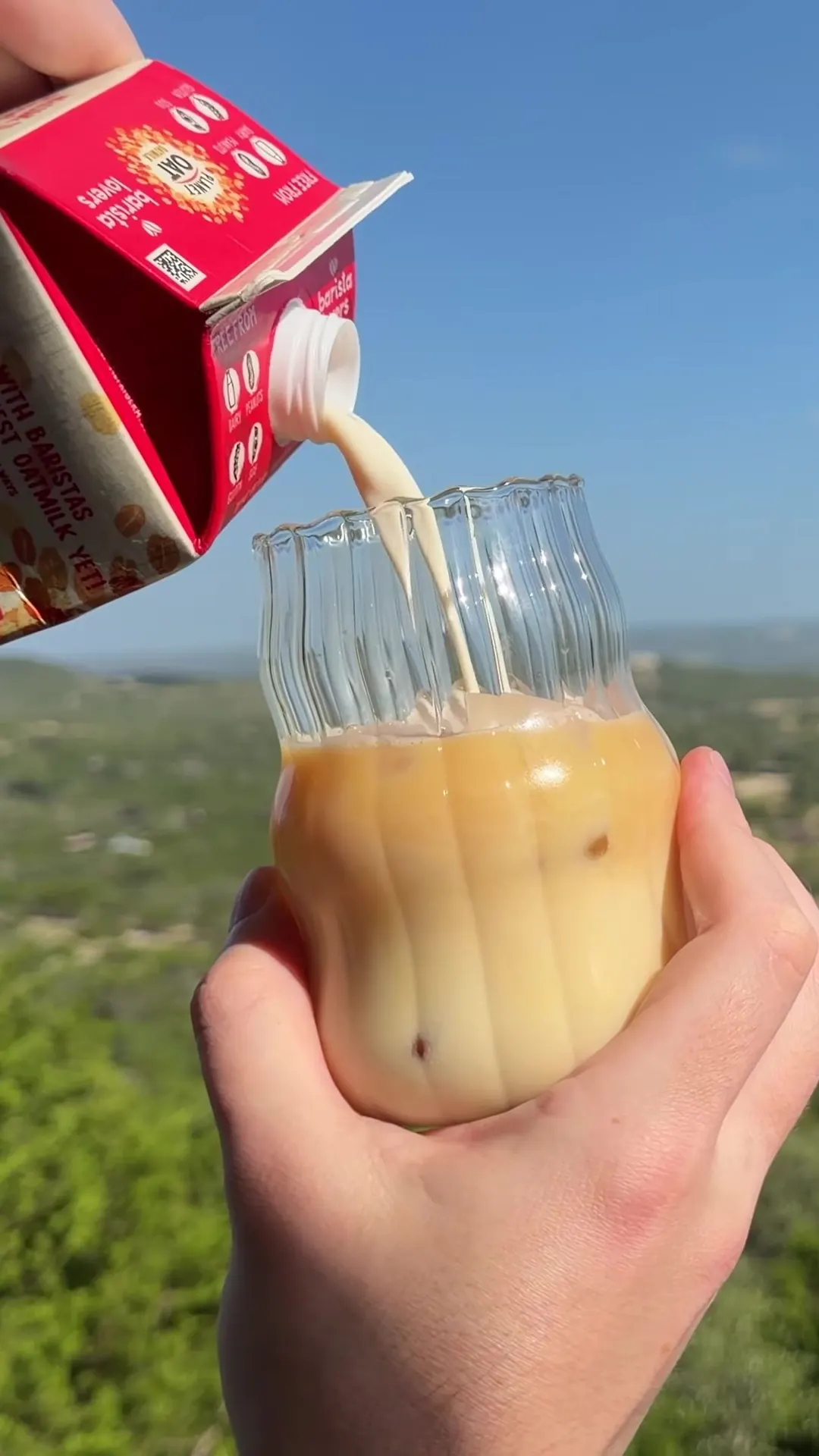 A hand pouring milk into a glass of iced coffee, held up against a wide green valley under a blue sky