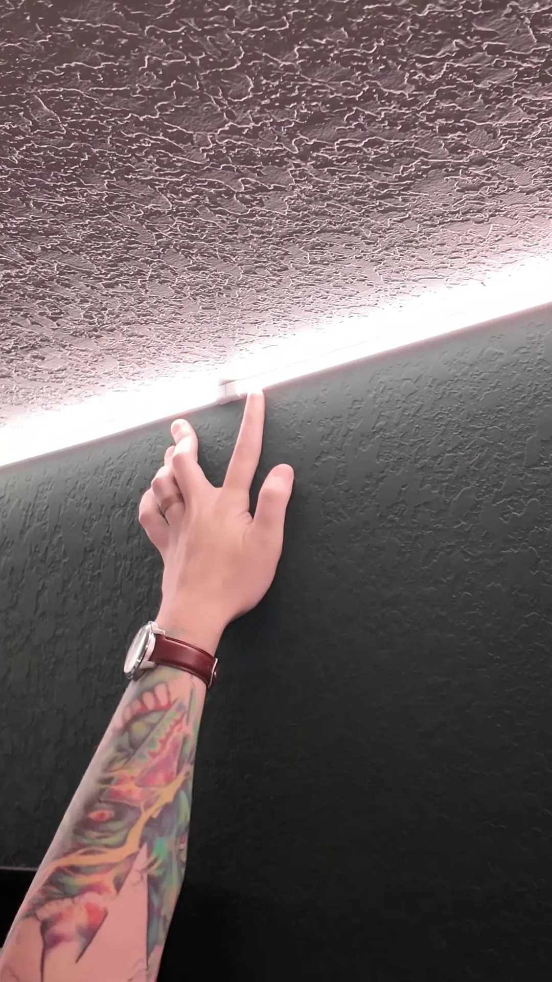 A tattooed arm reaching up to a glowing LED strip that runs along a dark wall below a textured ceiling