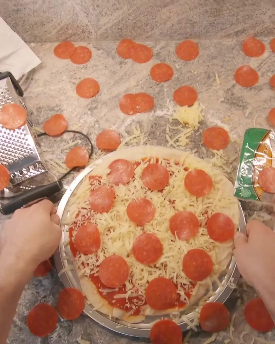 Overhead first-person view of hands on a pepperoni pizza on a stone counter, a grater beside it